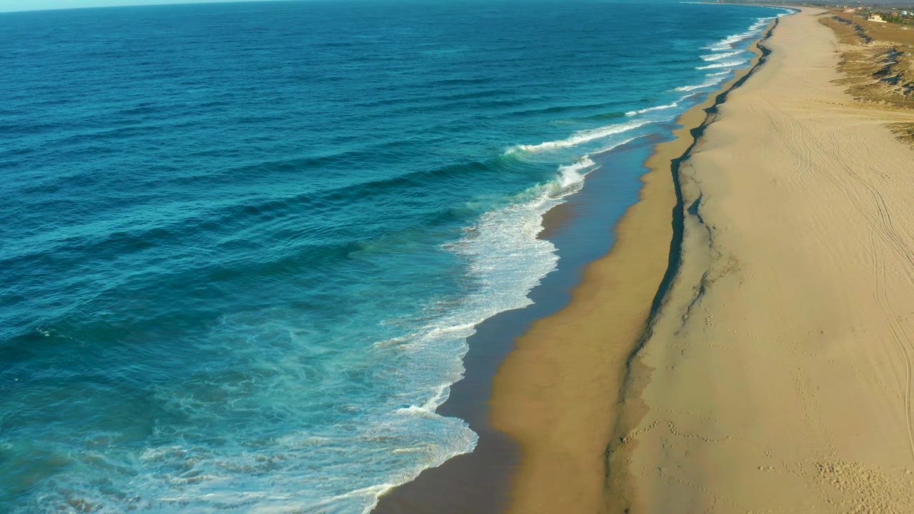 Aerial coastal frame from Todos Santos showing a broader section of turquoise shallows, white wave lines, a wide beige beach, a dark receding-water channel, and no visible people.