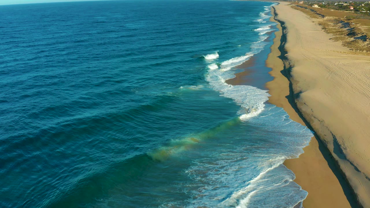 A second aerial frame over Todos Santos shows a bright white wave breaking farther along the diagonal beach, with open sand and no visible people.