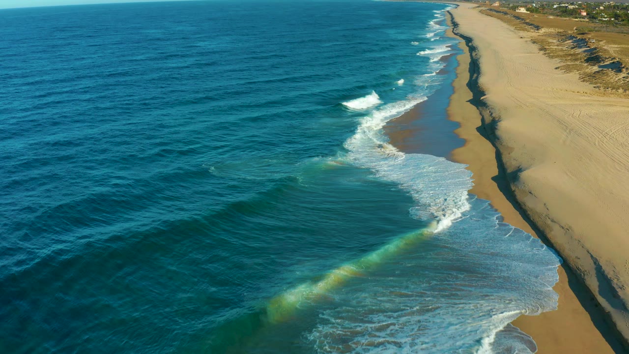 Aerial view in Todos Santos with darker blue water offshore, a pale green swell near the lower center, white foam at the beach edge, and no visible people.