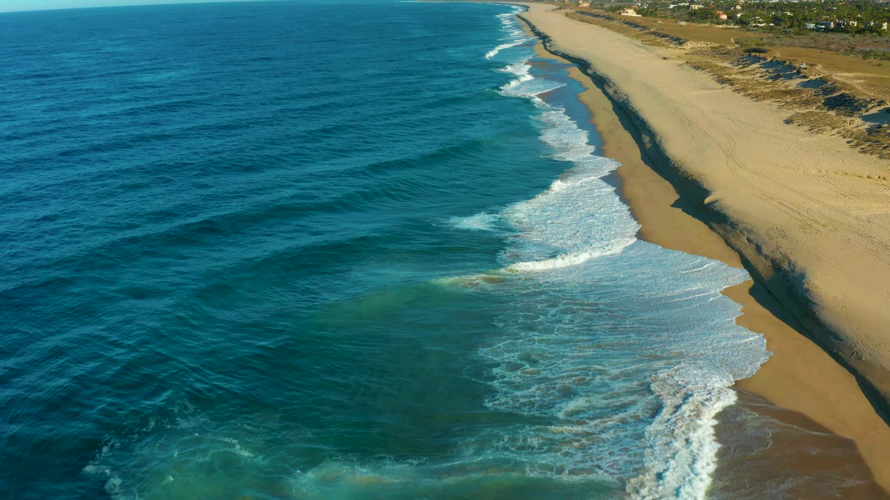 Aerial frame from Todos Santos showing a larger fan of white foam crossing the wet shoreline while blue water fills most of the left side and the empty beach runs into the distance.