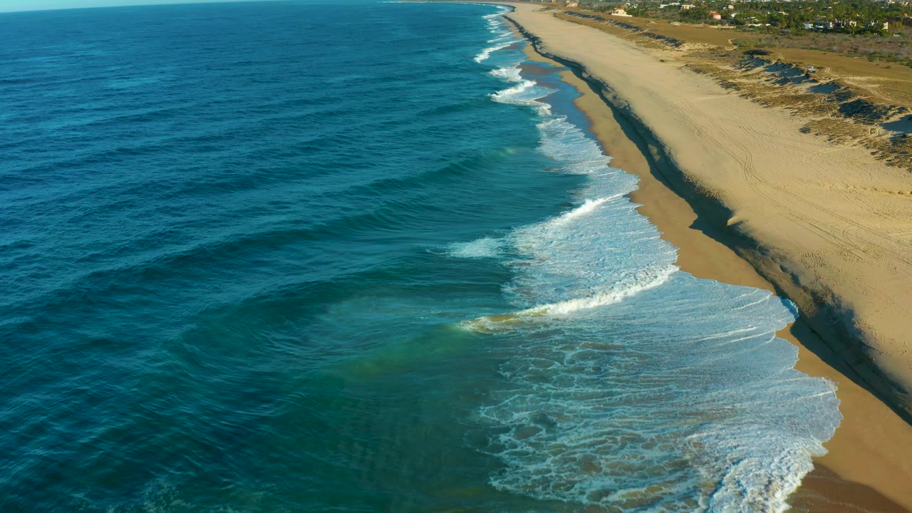 Aerial view in Todos Santos with darker offshore water, a curling surf line near the center, open tan beach to the right, and no visible people or vehicles.