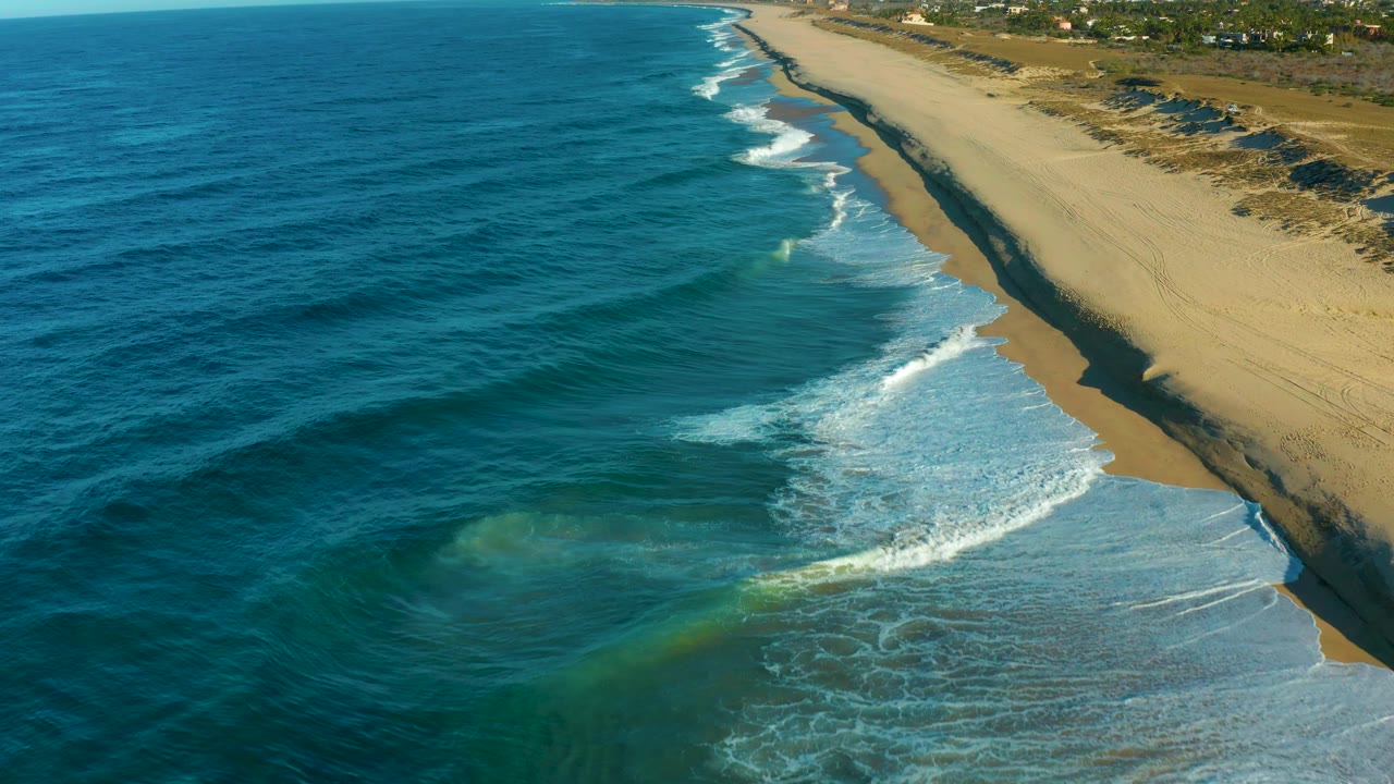 Todos Santos shoreline seen from above as white foam pushes diagonally from lower center toward the right-side beach, with turquoise shallows near the surf and no visible people.