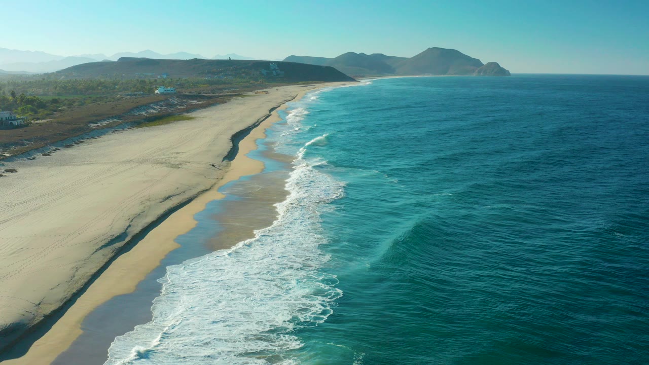 High aerial view over a Todos Santos sandy beach, with blue ocean on the right, white surf bands along the shore, distant hills on the horizon, and no visible people.