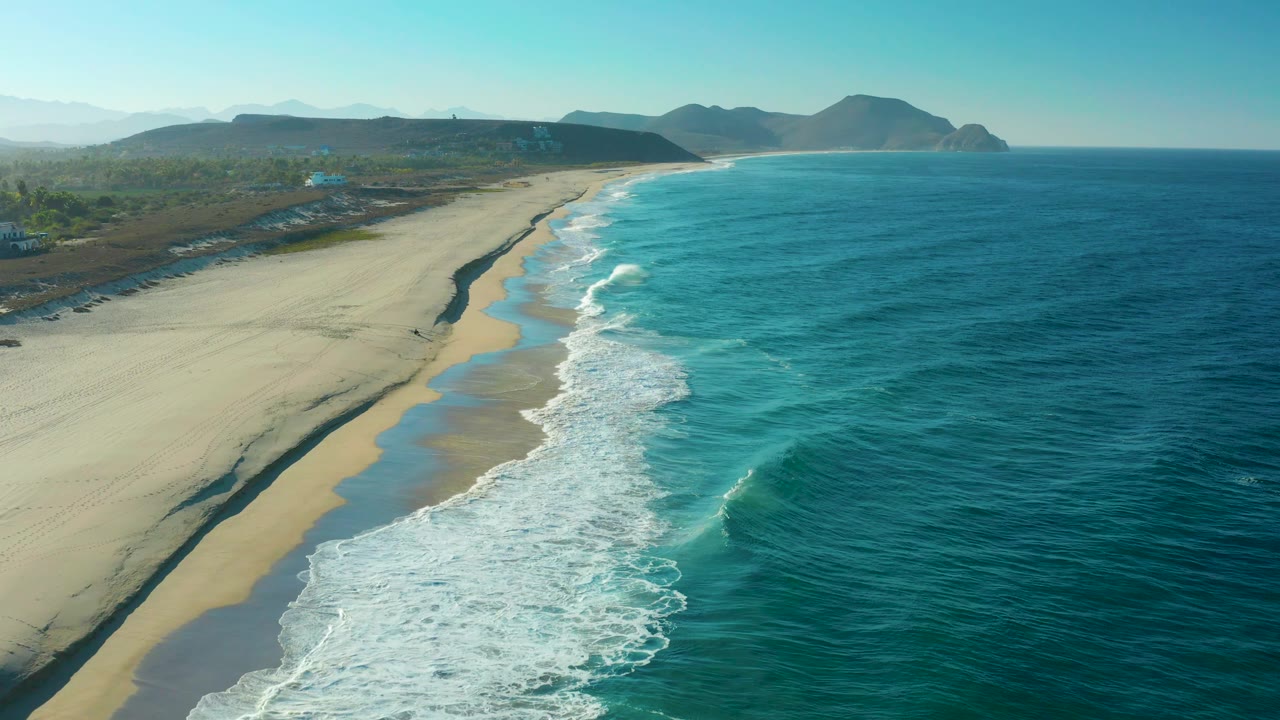 Aerial view of the Todos Santos shoreline with more open ocean visible to the right, white breakers tracing the beach edge, inland greenery beyond the sand, and no visible people.