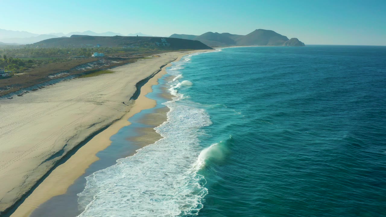 Aerial frame over Todos Santos showing a long pale beach, a dark wet-sand channel near the surf, turquoise water, low hills in the distance, and no visible people.