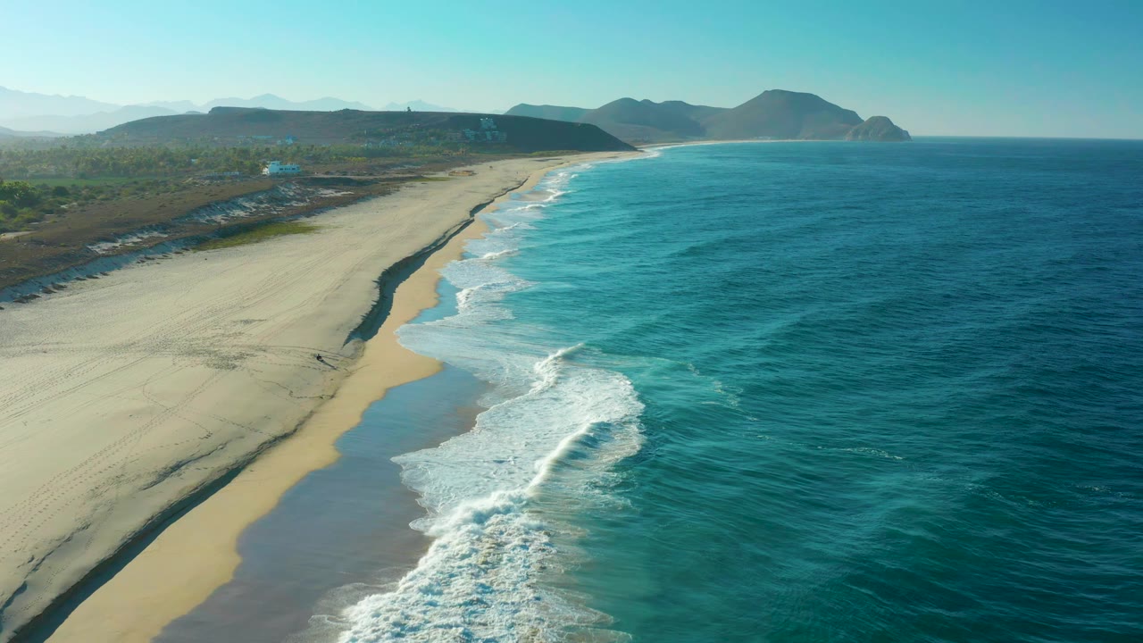 High coastal view in Todos Santos, Baja California Sur, showing a long sand strip, breaking white waves near the lower center, open blue water, distant hills, and no visible people.
