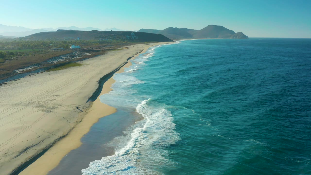 Oblique aerial frame over Todos Santos, Baja California Sur, with surf curling at the lower edge, a long pale beach to the left, deep blue ocean to the right, distant hills, and no visible people.