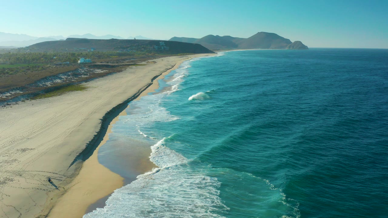 Aerial view of waves rolling onto a long sandy beach with mountains beyond.