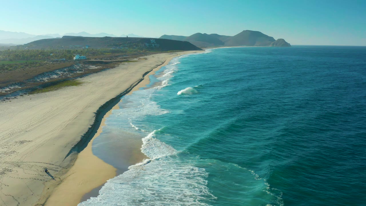 Aerial view of the Todos Santos shore one second into the clip, with a white wave breaking near the center, wet sand curving along the beach, and no visible people.