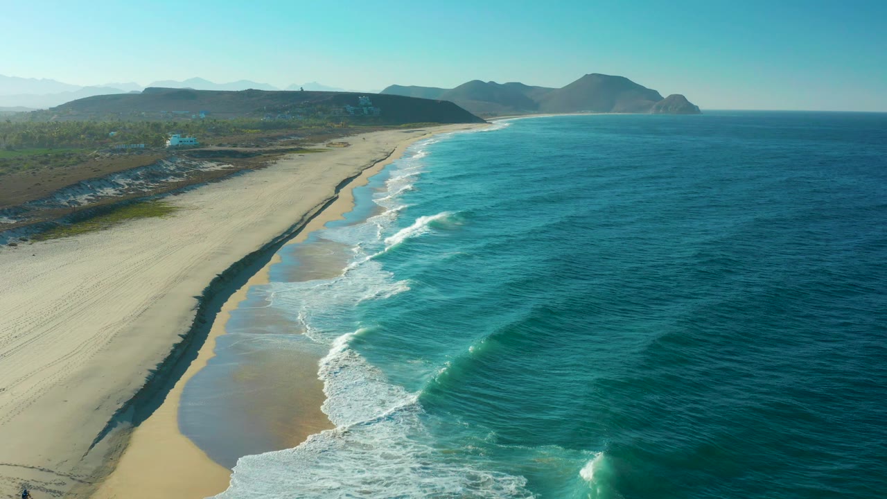 Mid-clip aerial view of the Todos Santos beach, with a broad wet-sand strip reflecting light, white foam along the shore, blue water to the right, and no visible people.