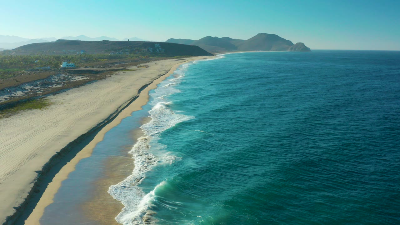 Aerial frame over Todos Santos, Baja California Sur with turquoise ocean on the right, pale beach on the left, distant mountains on the horizon, and no visible people.