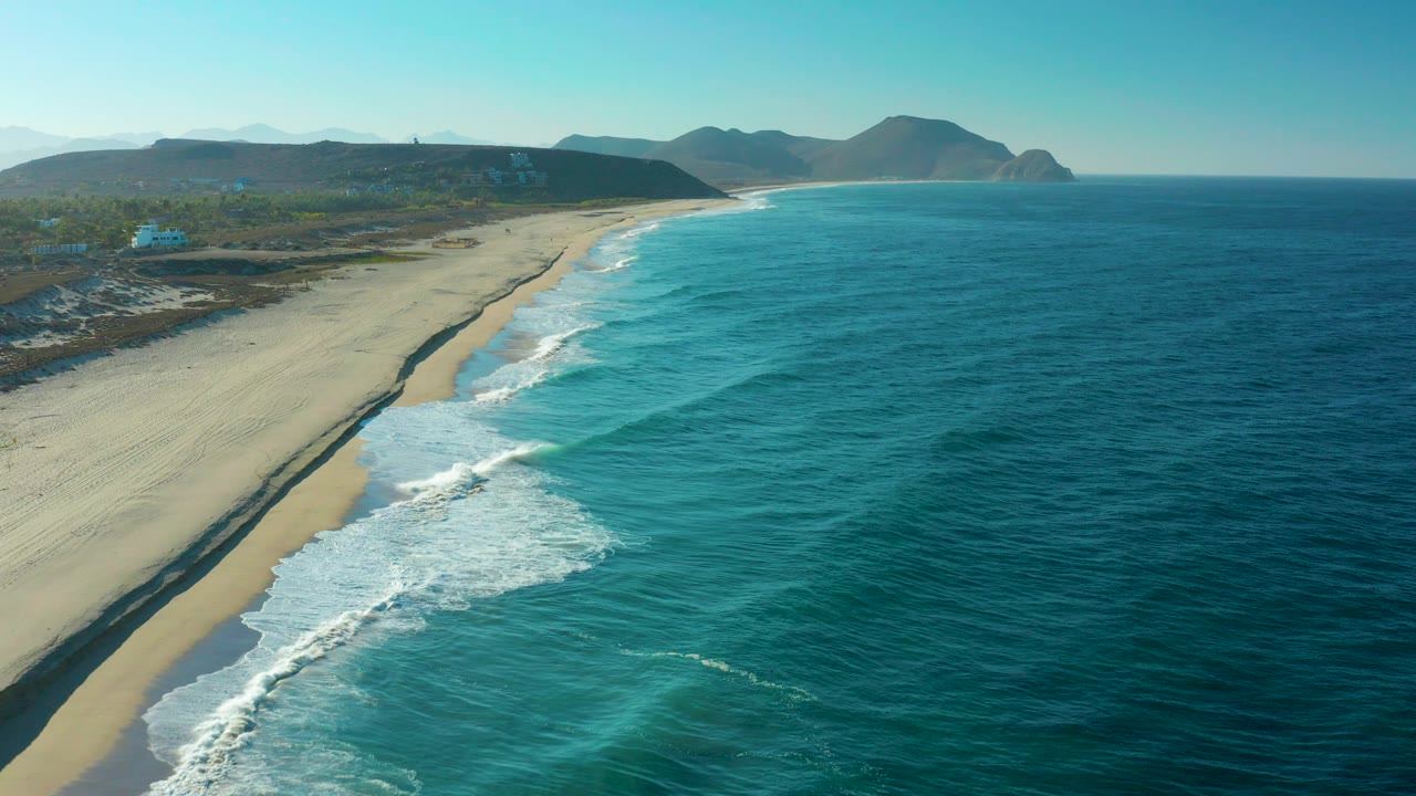 Aerial view over Todos Santos, Baja California Sur, showing turquoise ocean on the right, white surf on a sandy beach, distant hills, clear sky, and no visible people.