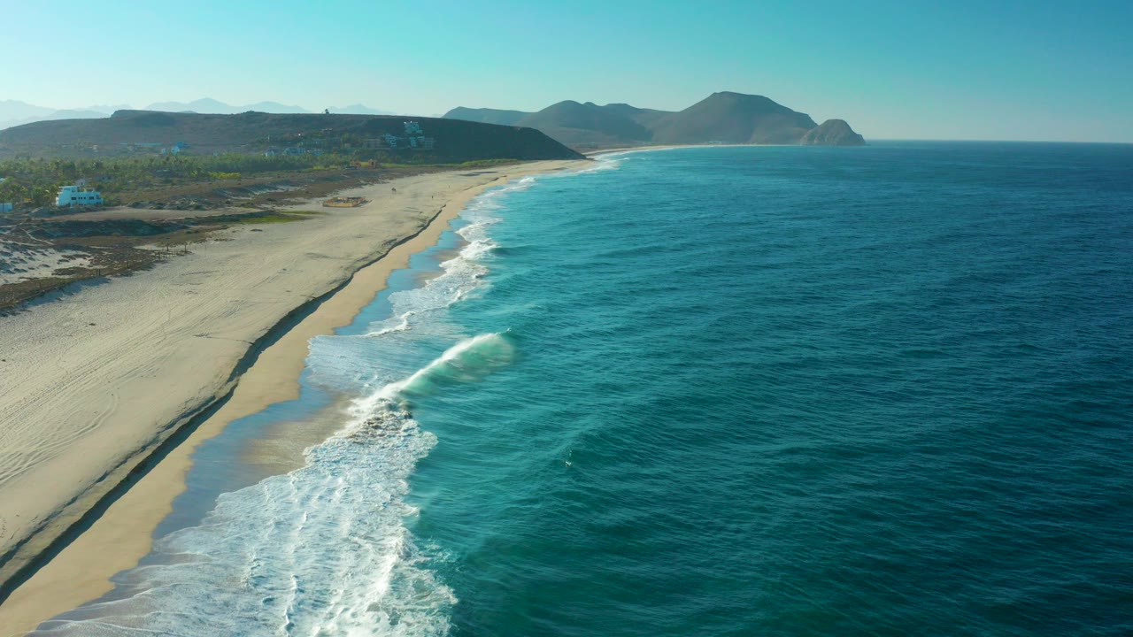 Aerial view of waves breaking along a sandy beach beside blue ocean.