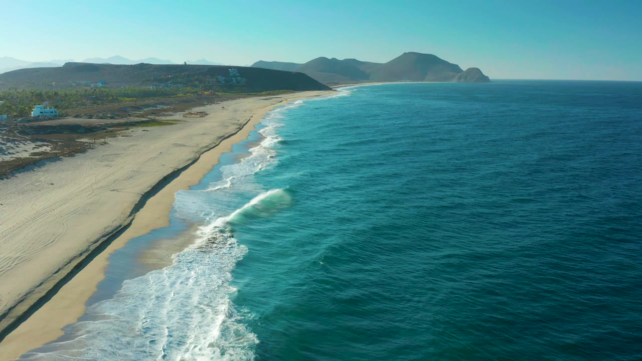 Aerial frame over the Todos Santos shoreline with turquoise water pushing white foam onto pale sand, dark coastal scrub inland, distant hills, and no visible people.