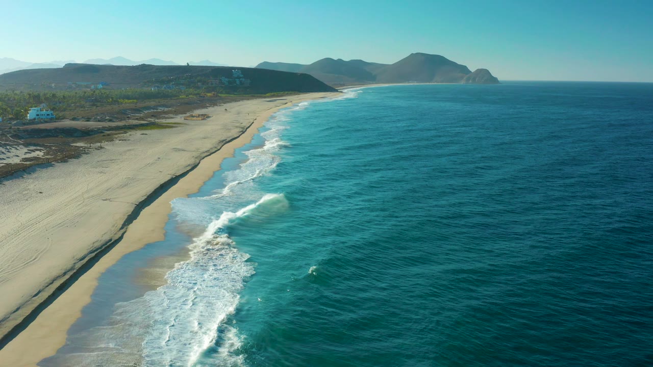 High coastal view in Todos Santos with darker blue water farther offshore, foamy wave sets near the sand, a low inland strip, distant hills, and no visible people.