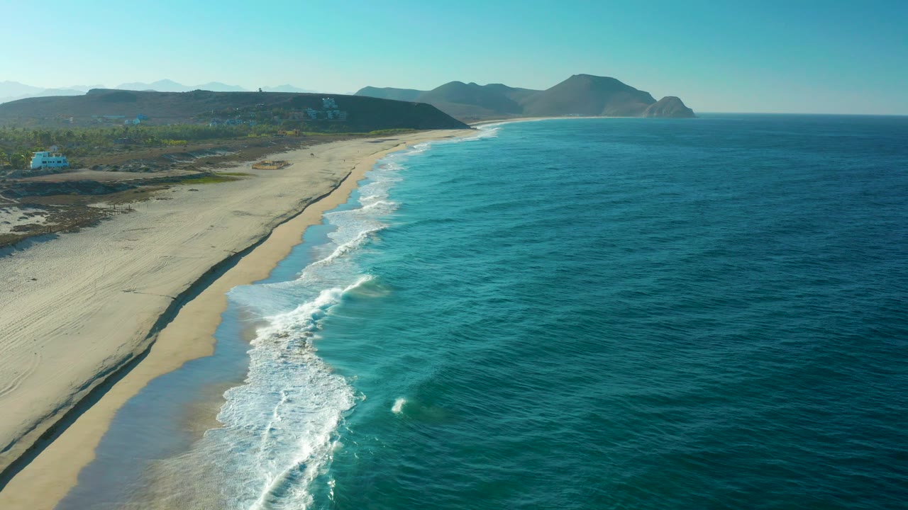 Aerial view of the Todos Santos beach as a wide band of sand runs along the left, blue ocean fills the right, surf curls near the lower edge, and no visible people appear.