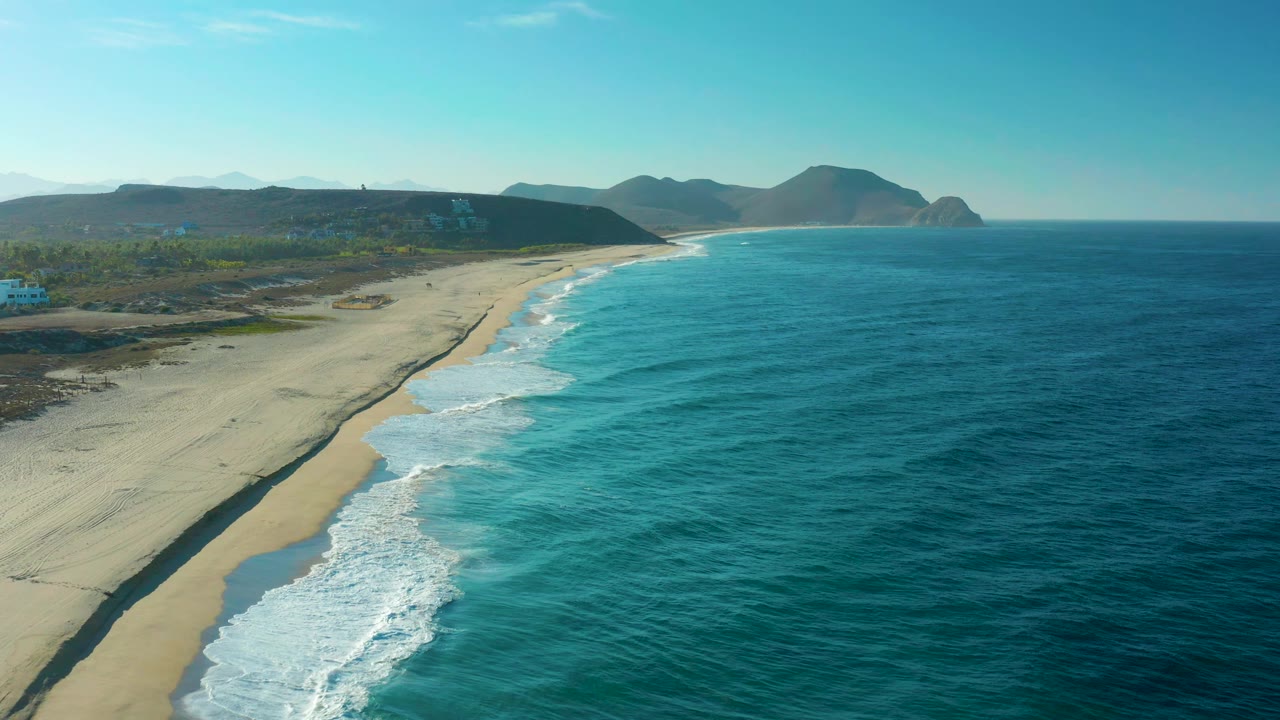 Aerial frame from Todos Santos, Baja California Sur showing white wave foam advancing along the sandy beach, blue-green water offshore, low hills in the distance, and no visible people.