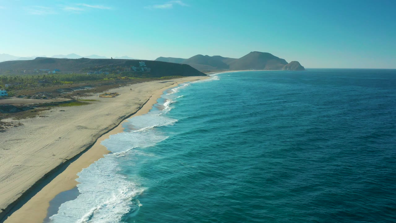 Wide aerial frame over Todos Santos, Baja California Sur with the sandy beach running diagonally, rolling surf at lower left, coastal vegetation inland, distant hills, and no visible people.