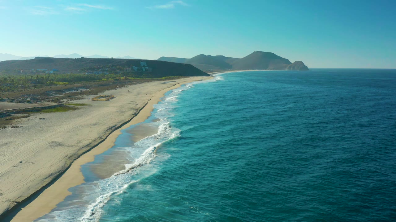 Elevated Todos Santos beach view with white wave foam advancing along the curved shoreline, inland scrub beyond the sand, and no visible people.