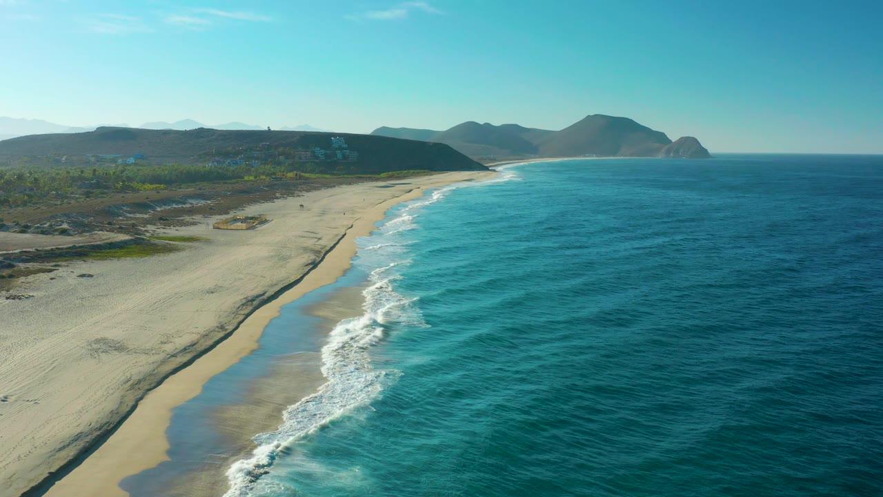 High drone-like view over Todos Santos beach, where white surf curls onto empty sand below coastal vegetation and low hills, with no visible people.