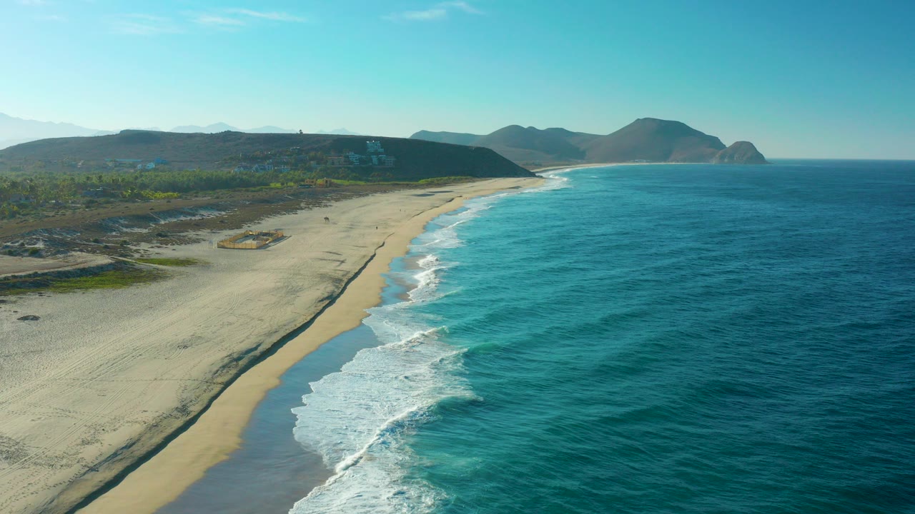 Aerial view of sandy beach, turquoise ocean, surf, and distant hills.