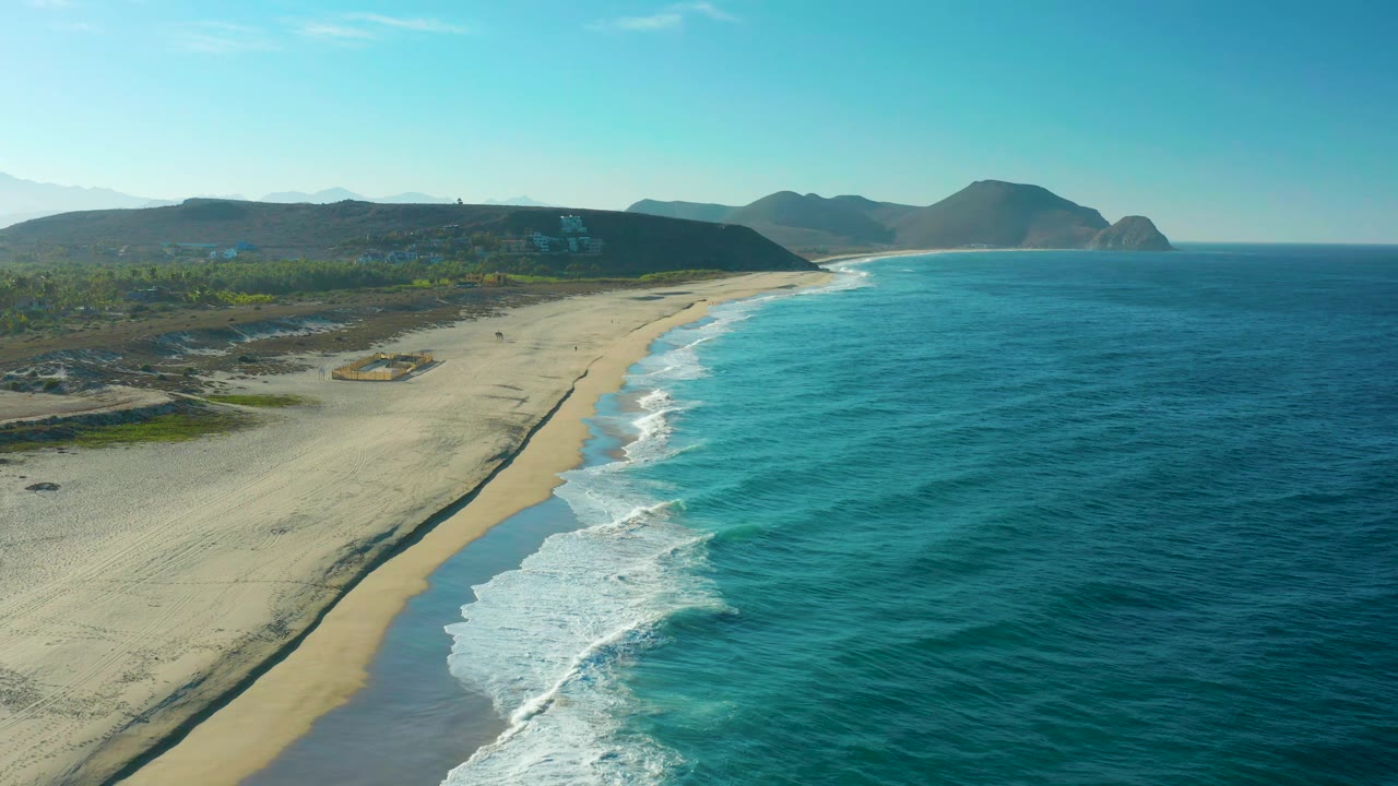 Aerial frame in Todos Santos showing the surf line running diagonally along pale sand, open turquoise water filling the right side, distant hills on the horizon, and no visible people.