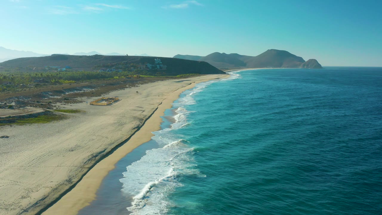 Aerial view of Todos Santos coastline with more open water on the right, a pale beach curving into the distance, low green inland terrain, distant hills, and no visible people.