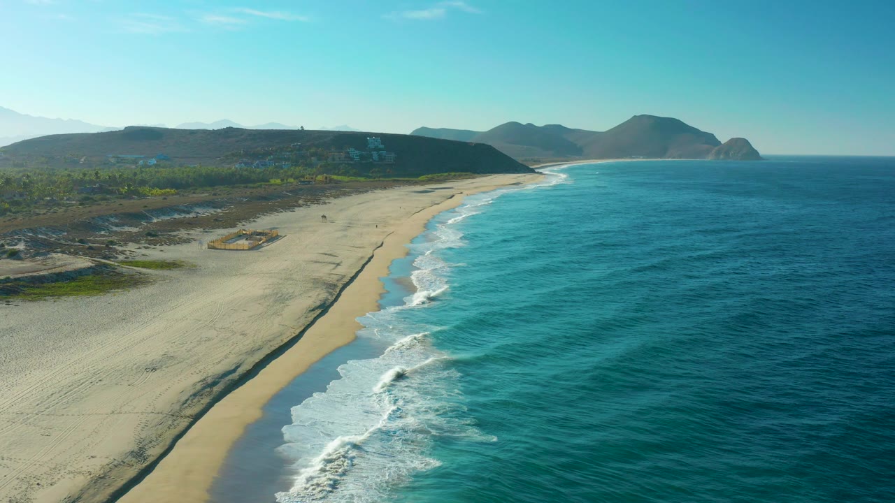 Aerial shot over Todos Santos with the sandy beach crossing the lower frame, textured turquoise water beside it, coastal vegetation inland, distant hills, and no visible people.