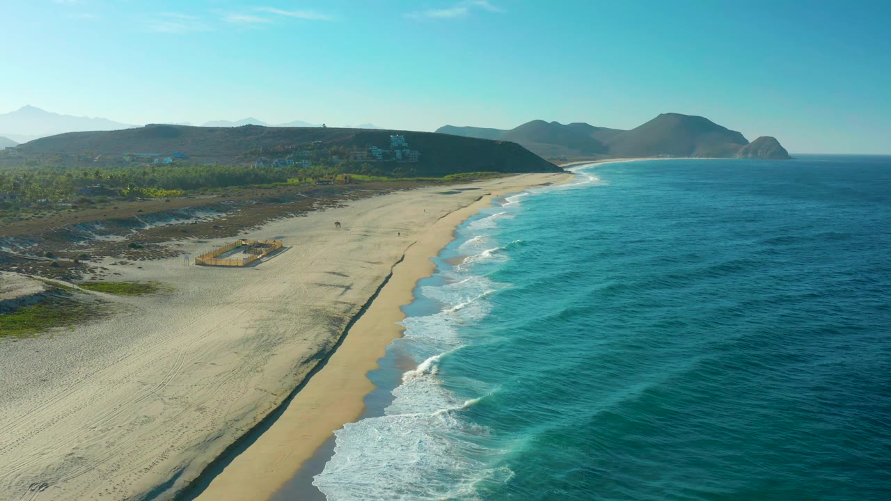 Aerial view in Todos Santos with the surf line running diagonally along the beach, turquoise water filling the right side, hills in the distance, and no visible people.