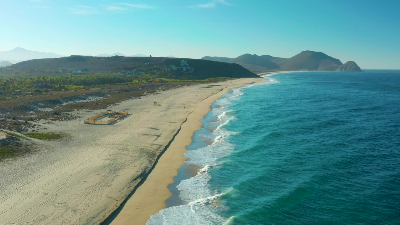 Aerial view over Todos Santos coastline with more inland scrub visible at left, a sandy beach curving toward distant hills, blue ocean at right, and no visible people.