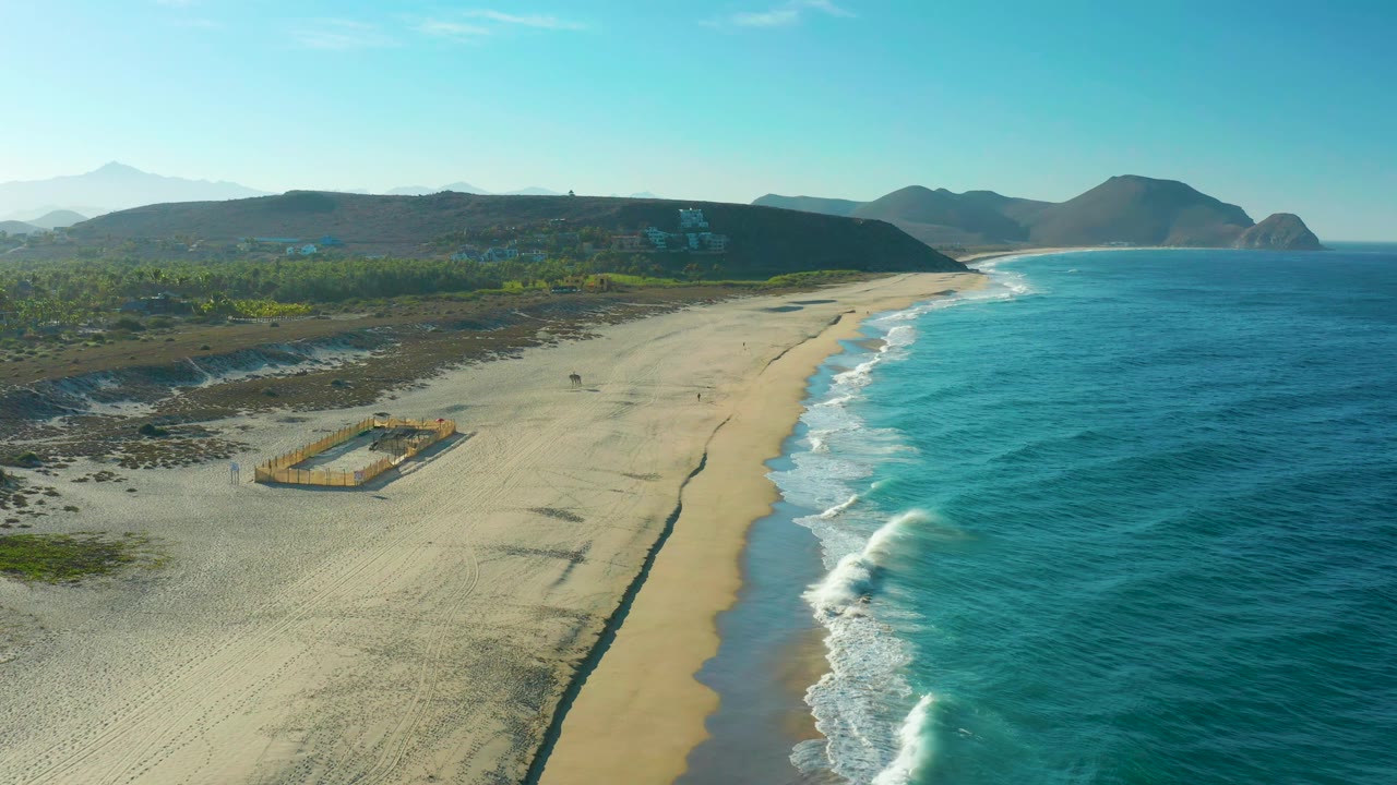 Aerial view over the Todos Santos shoreline as small waves reach the sand, with a rectangular fenced structure on the beach, scrubby inland ground, distant hills, and no visible people.
