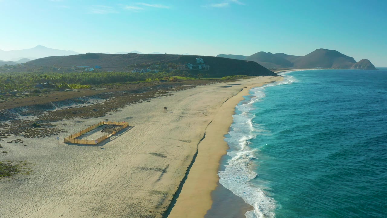 Mid-clip aerial view in Todos Santos with a fenced rectangular area near the lower left beach, foamy waves along the right shoreline, low green vegetation inland, distant hills, and no visible people.