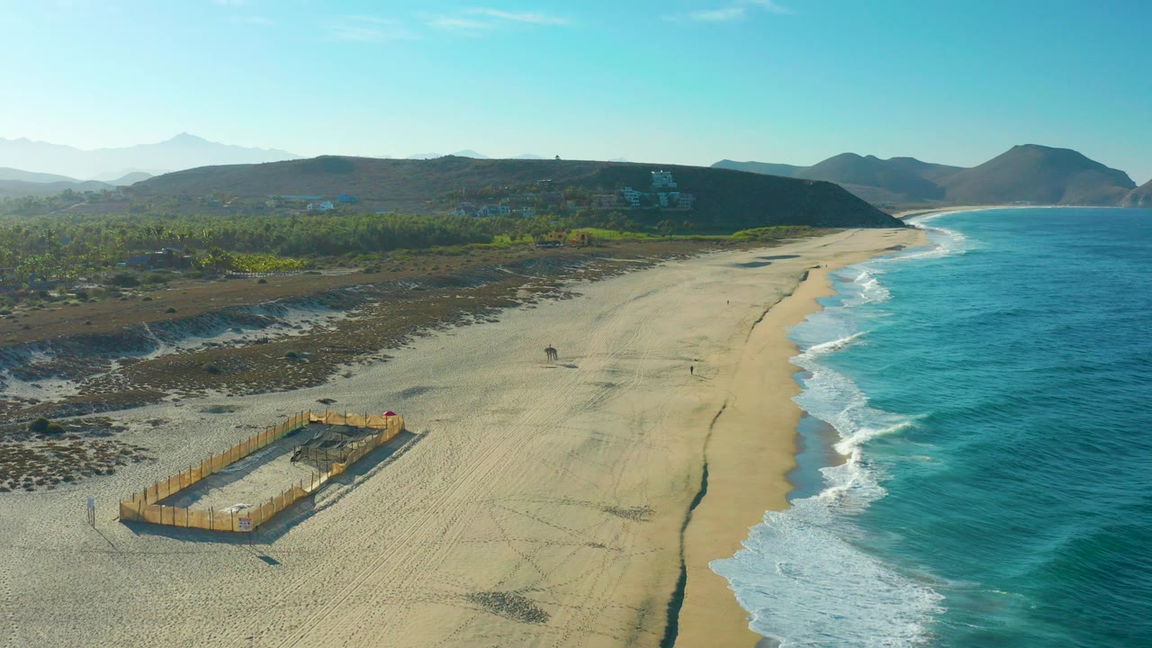 Aerial frame over Todos Santos beach with the wooden sand pit prominent at lower left, blue surf on the right, distant hills, and tiny walkers on the sand.