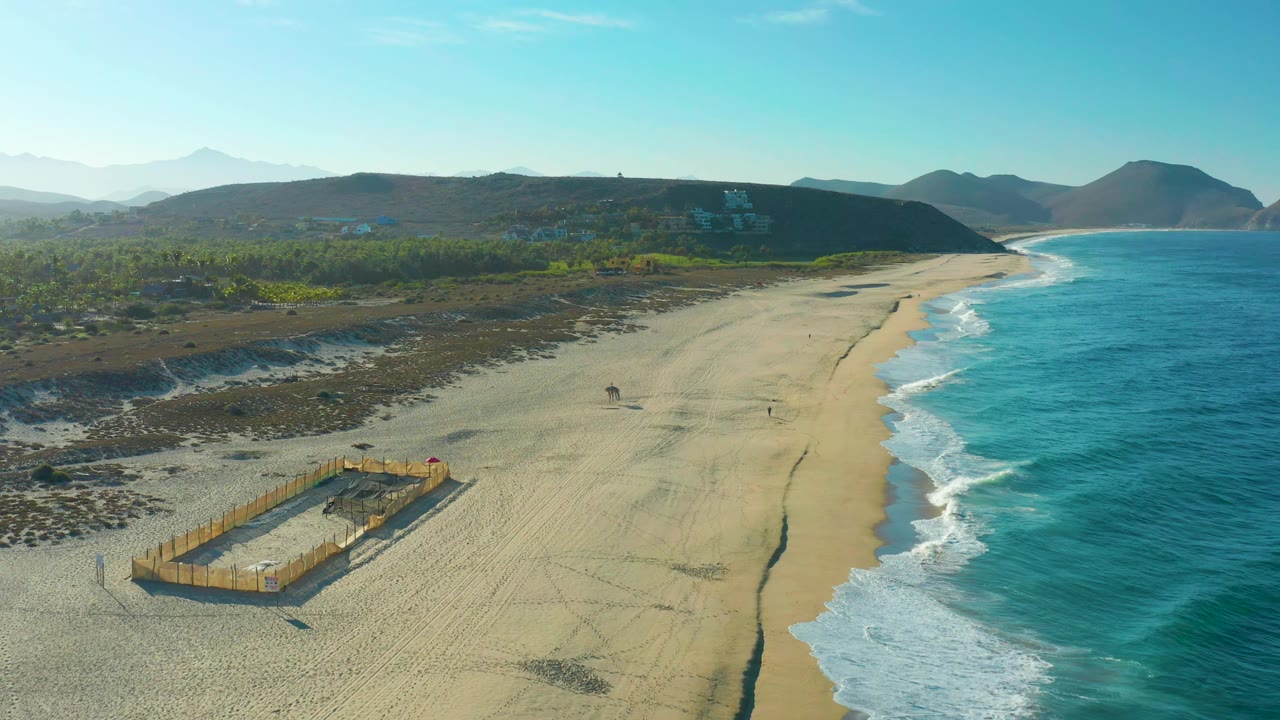 Drone view of shoreline with ocean waves, sand pit fencing, greenery, and hills.