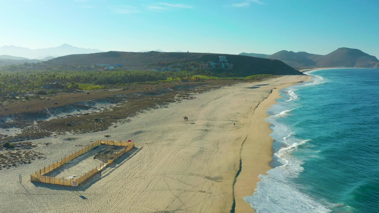 Wide drone frame over Todos Santos beach with turquoise water at right, the wooden sand pit lower left, a long empty stretch of sand, and tiny figures near the shoreline.
