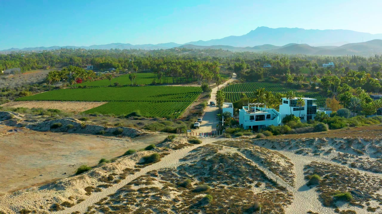 Aerial frame from Todos Santos, Baja California Sur showing a dirt road leading past dry ground toward green fields, a white building, distant mountains, and no visible people.