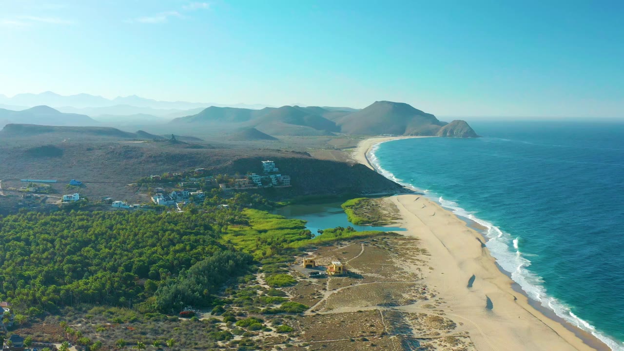 Aerial frame in Todos Santos showing the beach and surf line on the right, bright green vegetation and lagoon-like water inland, distant mountains, and no visible people.