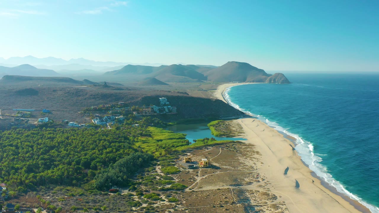 Elevated Todos Santos coastal view with the curving sandy beach below, ocean surf to the right, green fields and small buildings to the left, mountain horizon, and no visible people.