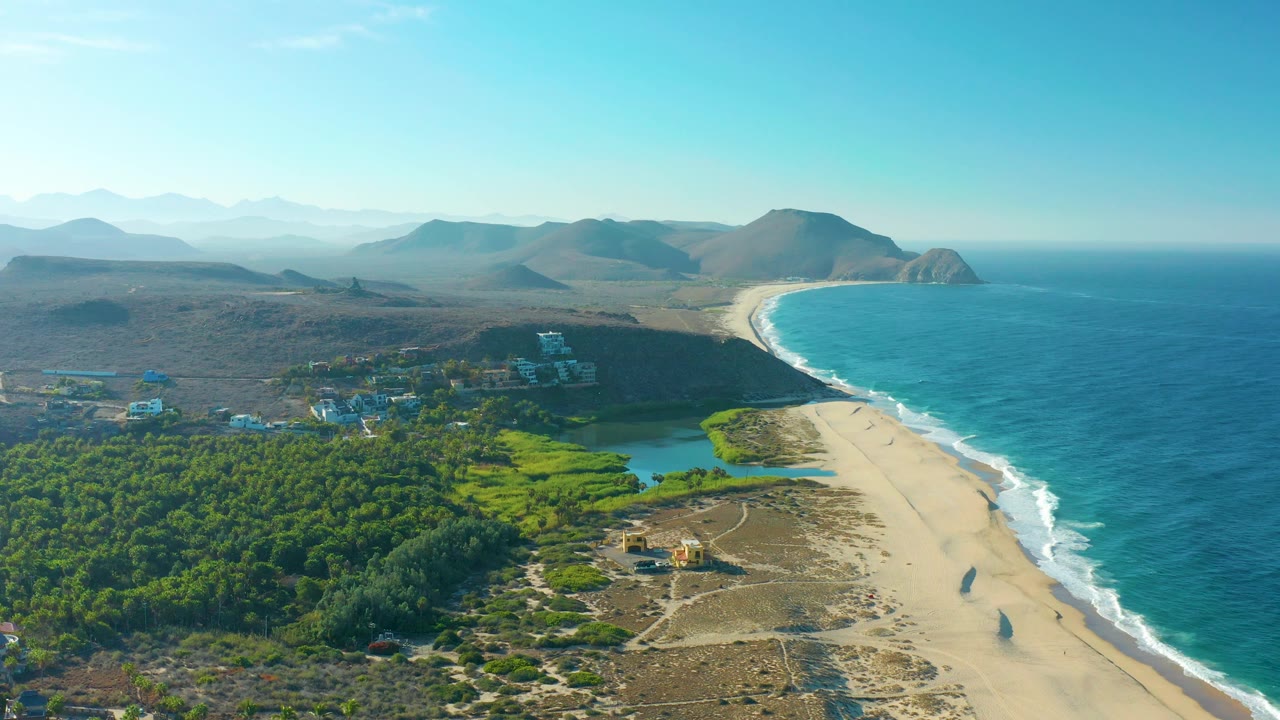 Aerial coastline with blue water, pale sand, inland greenery, dry hills, and clear sky.
