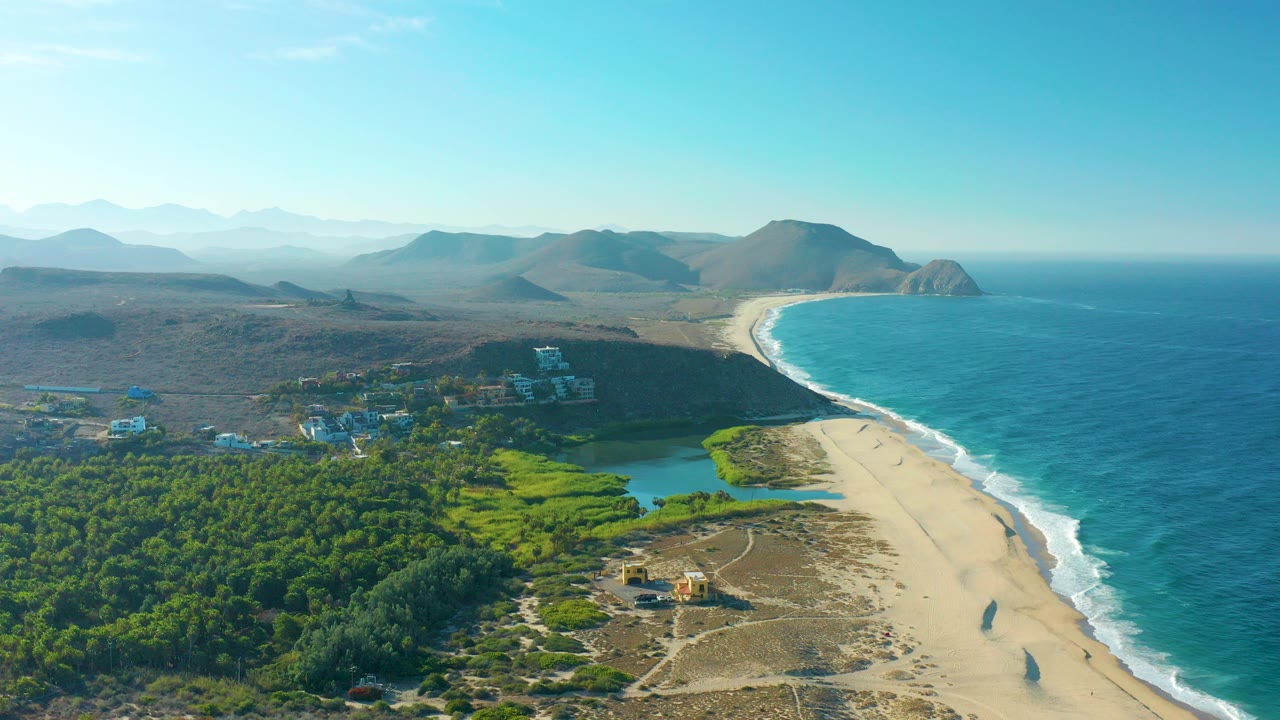 Opening aerial frame over Todos Santos, Baja California Sur, with blue ocean surf at right, a long sandy beach, green lagoon vegetation, scattered buildings, dry hills, distant mountains, and no visible people.