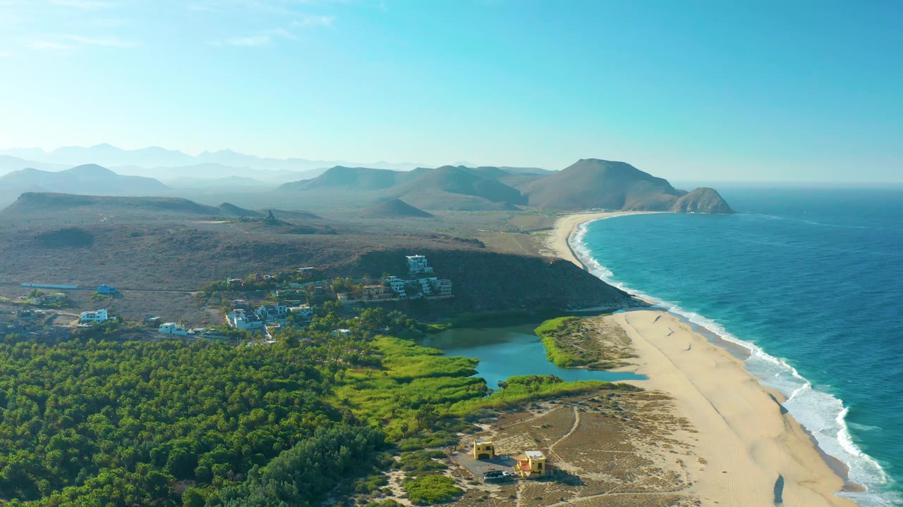 One second into the clip, the aerial view holds on the Todos Santos coastline with the lagoon and forest lower in frame, surf tracing the beach, and no visible people.