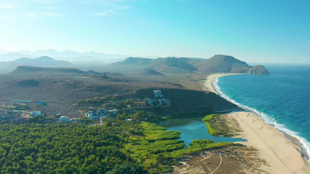 Opening aerial frame over Todos Santos, Baja California Sur, with green vegetation and lagoon-like water in the foreground, white coastal buildings, a curved sandy beach, blue ocean, distant mountains, and no visible people.