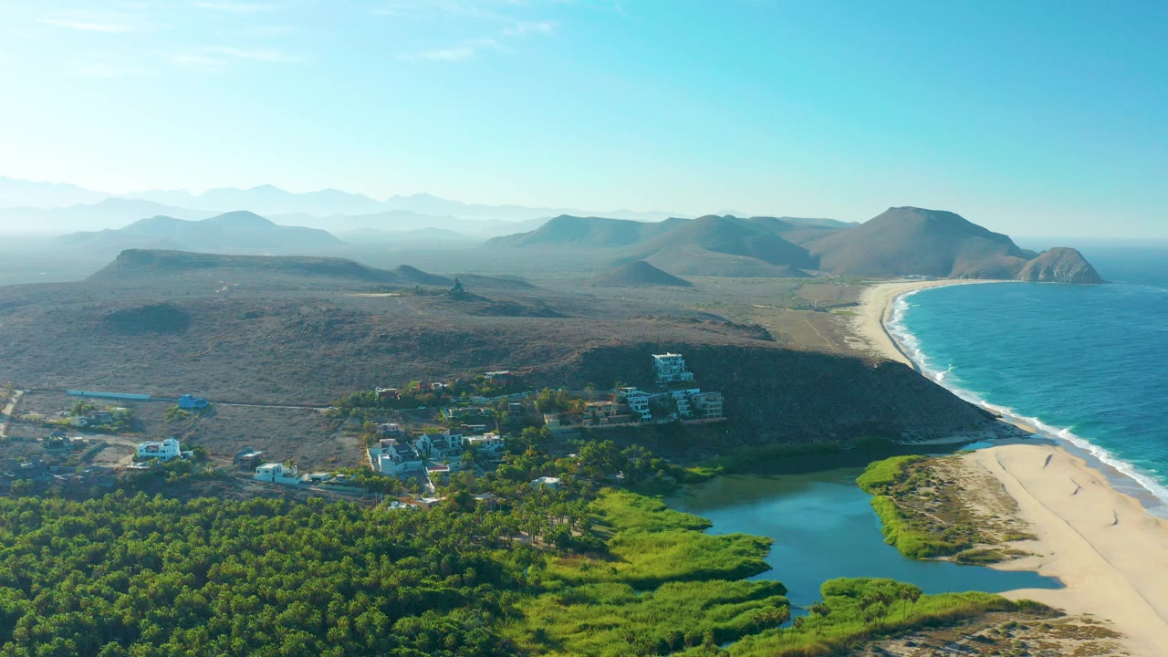 Aerial view in Todos Santos, Baja California Sur, showing trees and lagoon water in the foreground, a sandy beach and blue ocean to the right, dry hills beyond, and tiny people visible on the sand.
