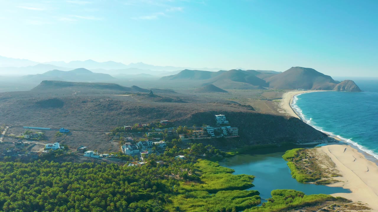High aerial view in Todos Santos with dry scrubland and hills across the background, the beach and ocean along the right edge, and lagoon water, trees, and low buildings below.