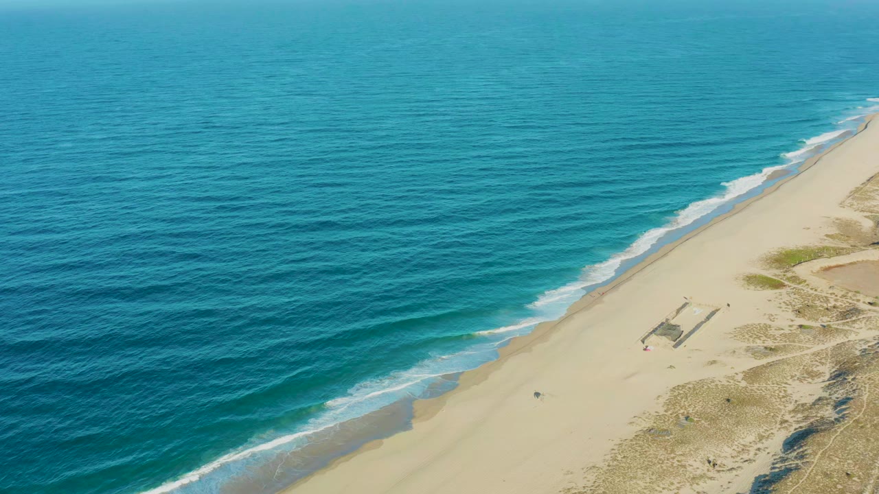 Aerial view over Todos Santos shoreline with deep blue water shifting to turquoise near the beach, a narrow white wave edge, pale sand, and no visible people.