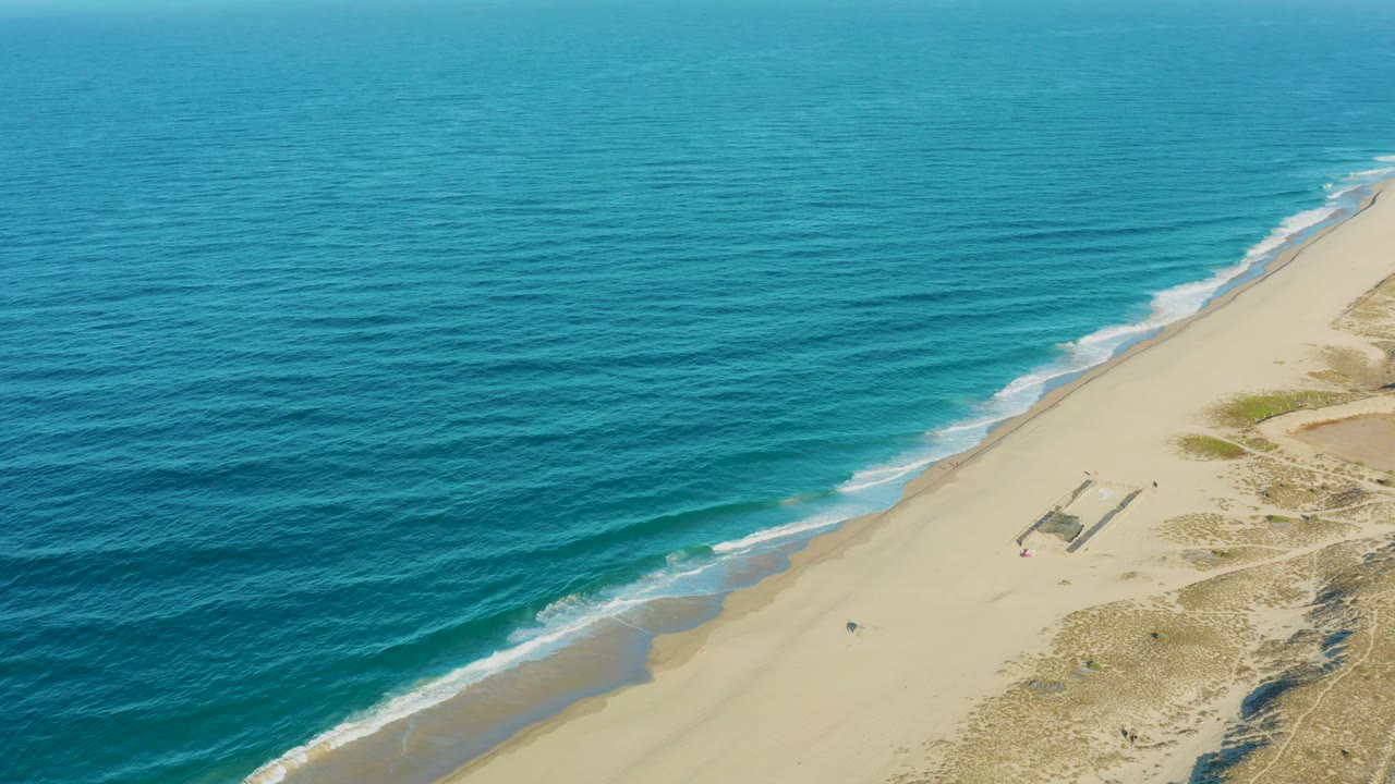 A wide aerial view in Todos Santos shows gentle surf tracing the beach, turquoise water filling most of the frame, dry dune vegetation to the right, and no visible people.