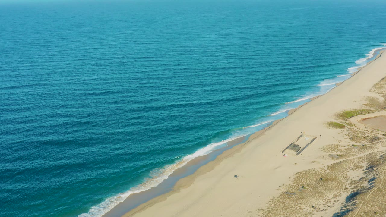 Aerial frame from Todos Santos, Baja California Sur showing blue ocean on the left, pale sand on the right, thin surf lines, dunes, and small beachgoers visible near the shore.