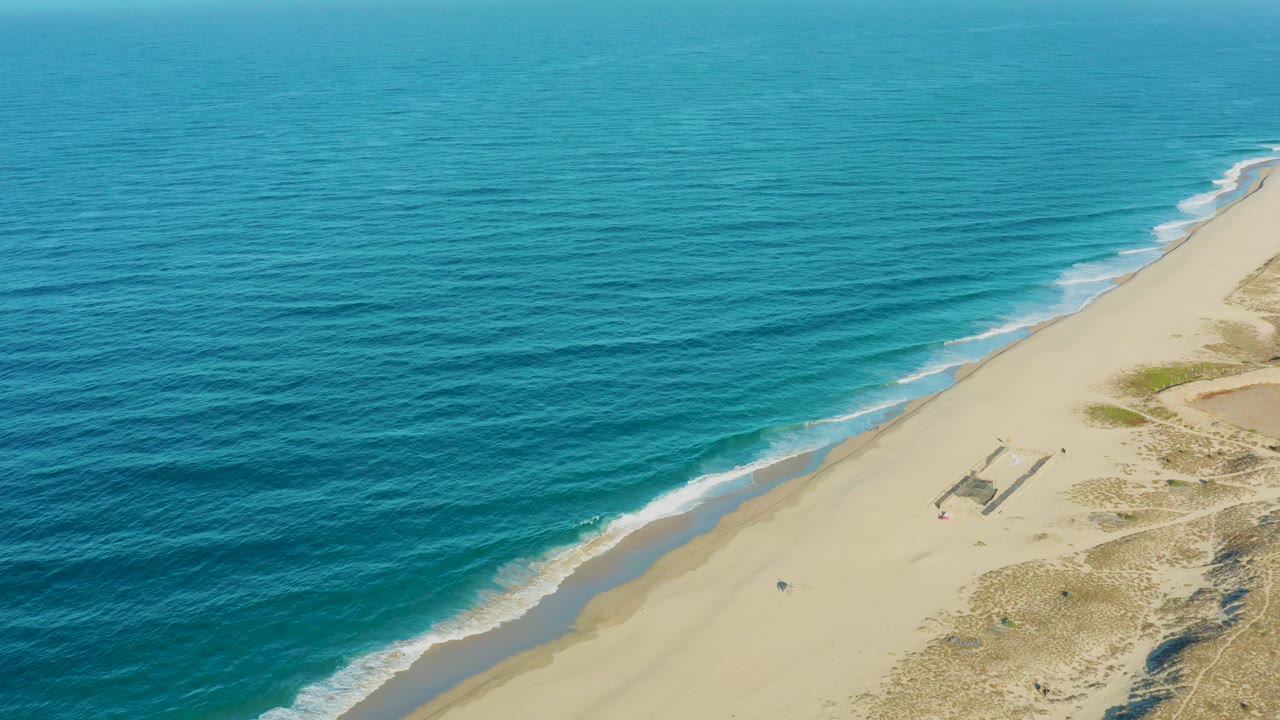 Blue ocean and sandy shoreline with surf bands and small figures on the beach.