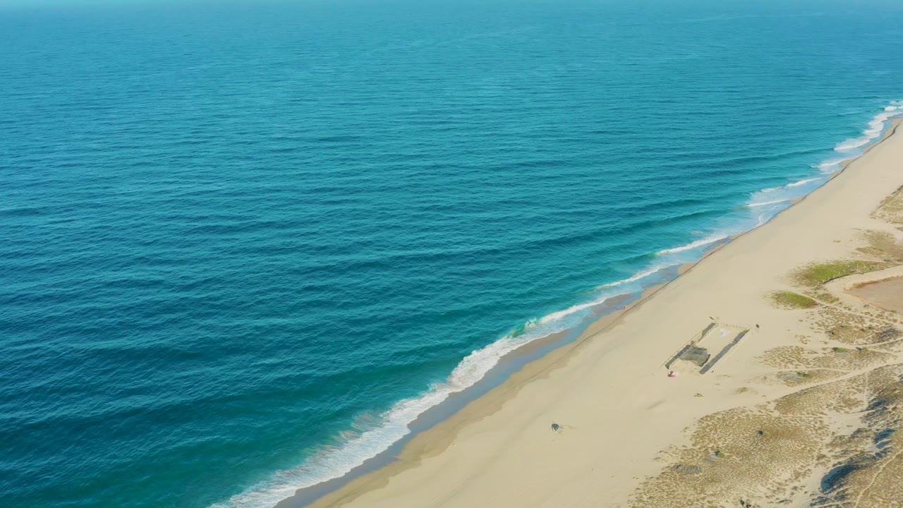 High aerial coastline view with blue water, pale beach, surf, and dunes.