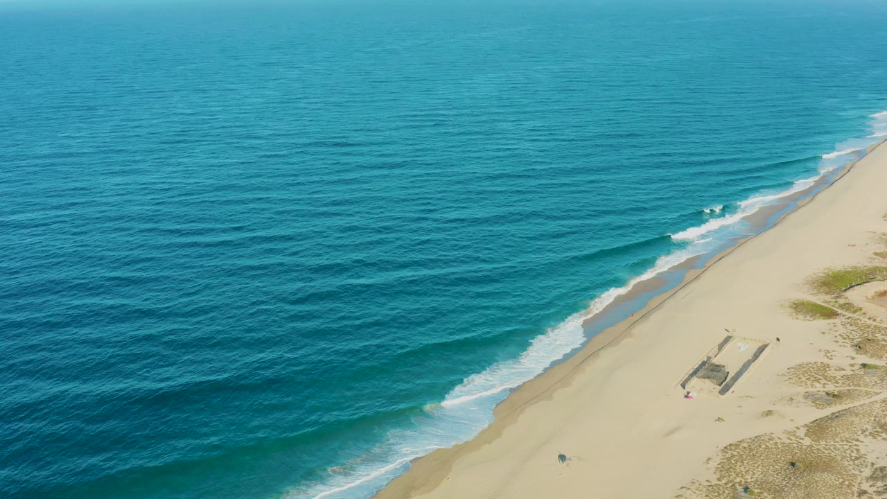Aerial view of a Todos Santos sandy shoreline with turquoise ocean on the left, white surf along the beach, and a small structure on the sand with a tiny person nearby.